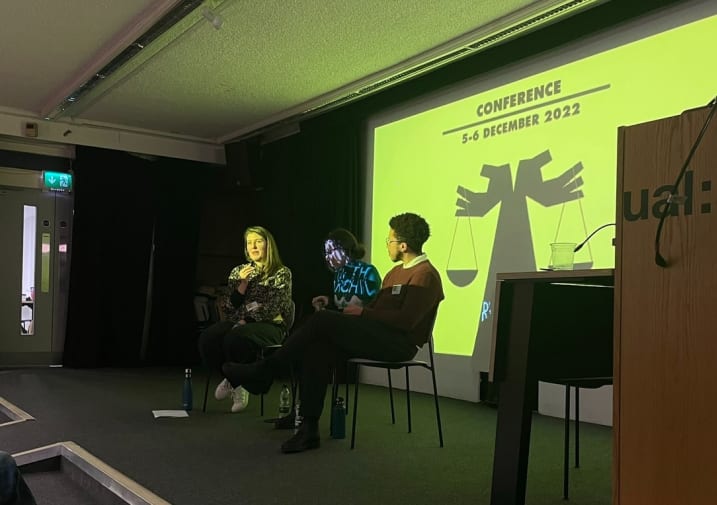Three people sit on a carpeted stage in front of a projected yellow screen and silhouette graphic of two hands holding weighing scales