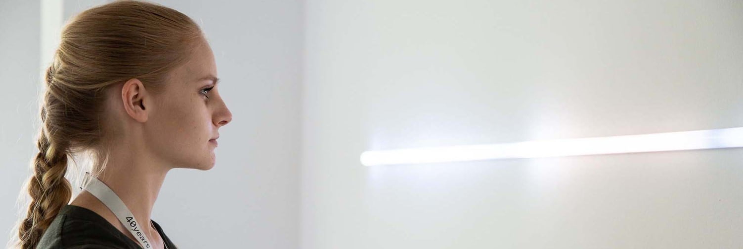 A woman in a white room looks towards a white neon tube light.