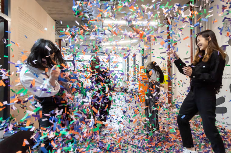 Four students throw green, orange, purple and white confetti at each other at the BA Graphic Design Summer Show at Camberwell