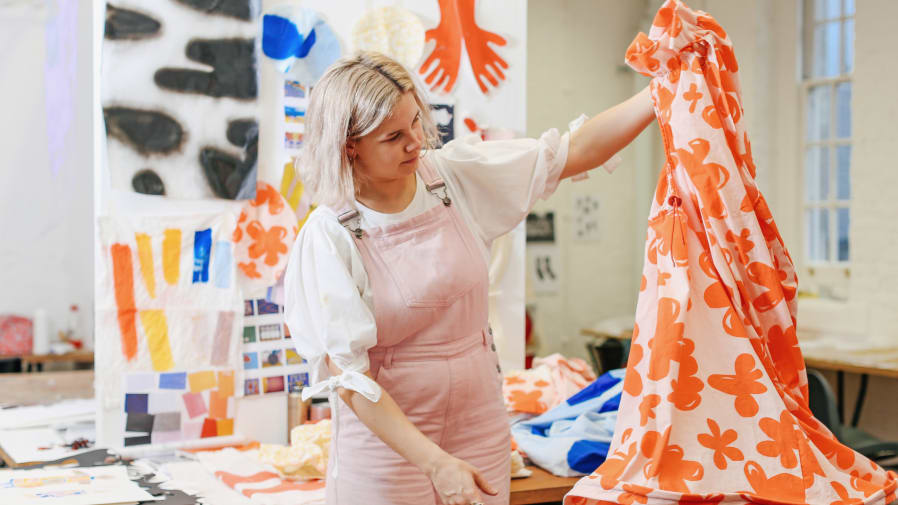 Person holding up orange and white patterned piece of fabric, with a backdrop of canvas with colourful paintings on.