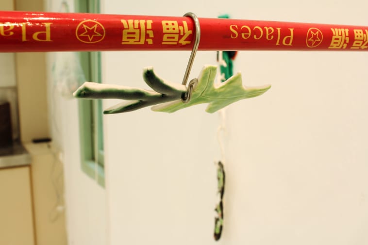 A detail photograph of an art work installation showing a red bar with yellow English and Chinese writing on it. Hanging from the bar is a silver butchers hook on which is a green ceramic object in the form of a tree or leaf.