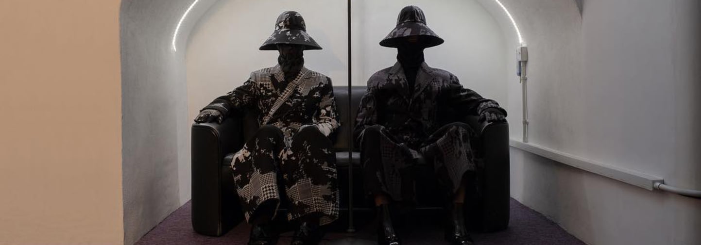 Two models wearing the same clothes with big hats and their faces covered, sitting side by side on two arm chairs