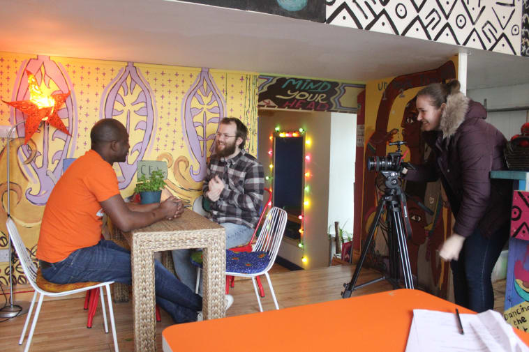 Two men, one client and one student, sit on a table in a cafe while a camera-woman films