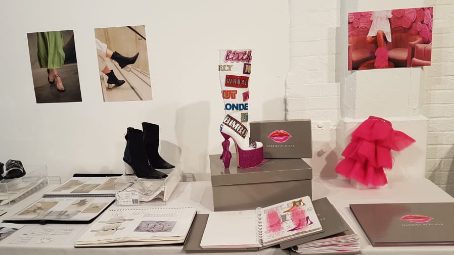 A black ankle boot with a concrete white heel stands next to a high heeled knee high boot in pink glitter which has badges on it with words like 'bitch' and 'bimbo.'