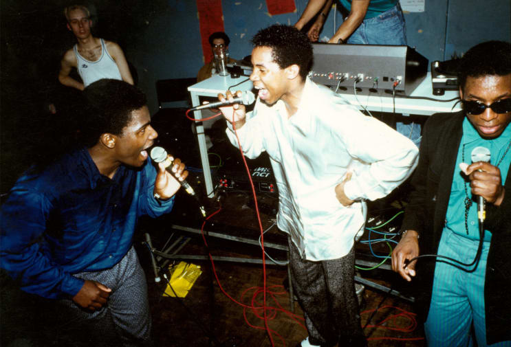 Three people standing together as a group singing into microphones in a club setting