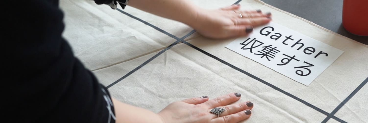 Two hands place a square of fabric which reads 'Gather' in English and Japanese onto a piece of cream cloth.