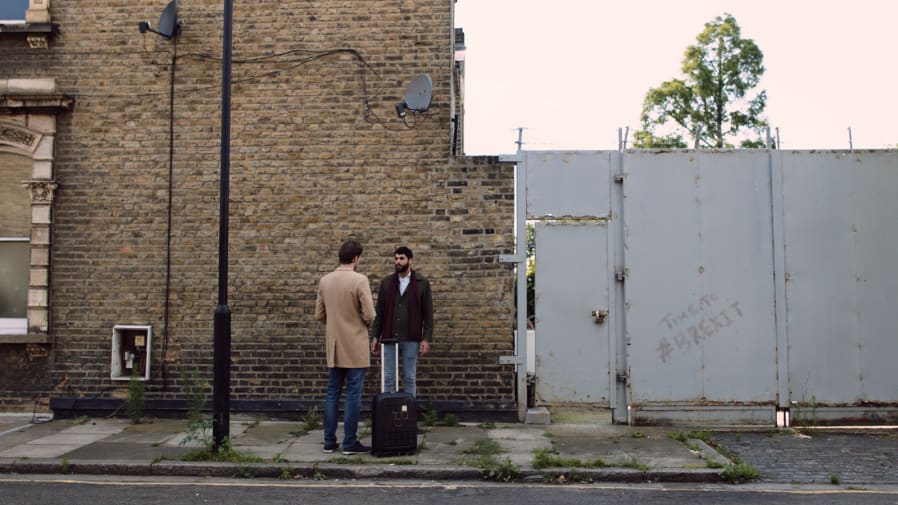 Two men stand and have a conversation in a residential street, one with a suitcase
