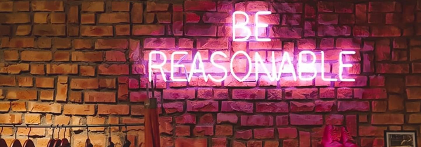 Be reasonable written in pink illuminous lights against a brick wall