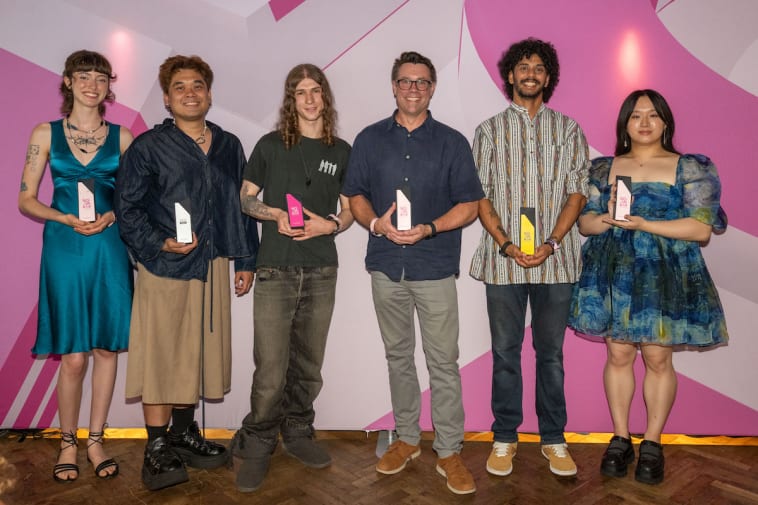 A group of student award winners stand in front of a pink background holding their awards