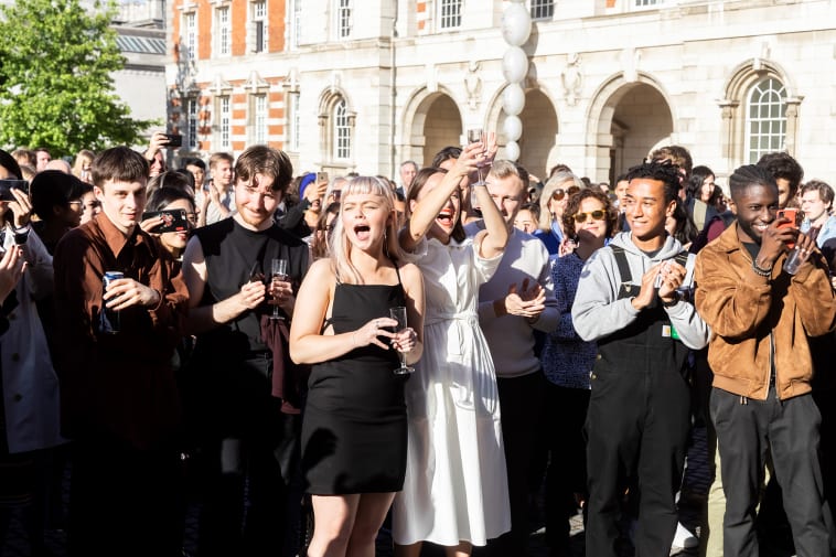 A group of Chelsea students stands in the Parade Ground at Chelsea cheering and shouting in celebration of a prize being awarded. They hold drinks and on the right a student is taking a photo with his mobile phone.
