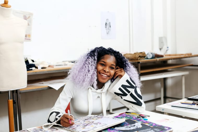 Woman with purple hair, smiling. Her sketchbook is open on the table
