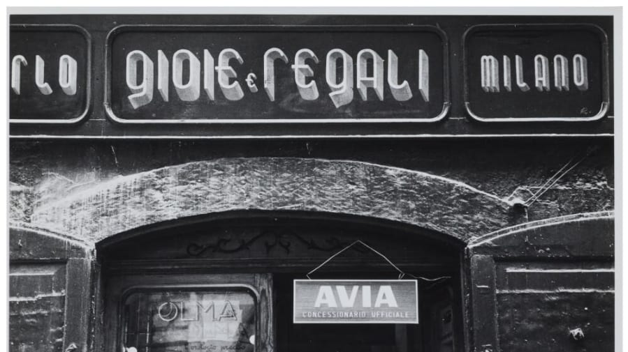 Black and white photograph of a shop sign with an elongated letter style, reading, ‘Gioie e Regali, Milano’.