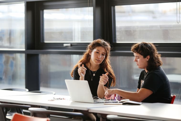 Two people sitting at a desk in front of a window with a laptop in front of them
