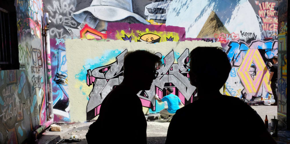 Two people watch a graffiti artist create work on an inner-city wall.