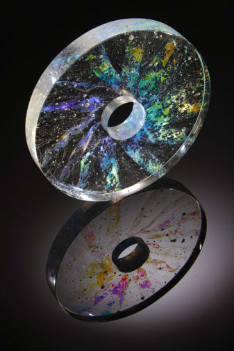 Glass circle with hole in the middle. Transparent glass with multi-coloured metalic slices radiating from the middle towards the edges and bubbles trapped inside.