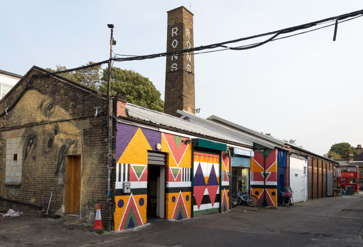 A row of low London premises covered in art on the front and side, shot from outside, with a bicycle in the doorway and a yard with a red bus just visible at the side