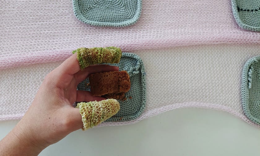 A knitted textile made with edible and inedible natural materials, and a hand wearing knitted finger wraps and making to pick up a piece of food to eat