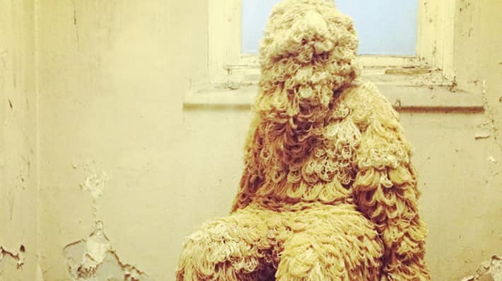 Image of a wooly human figure sat on a chair in a dilapidated room