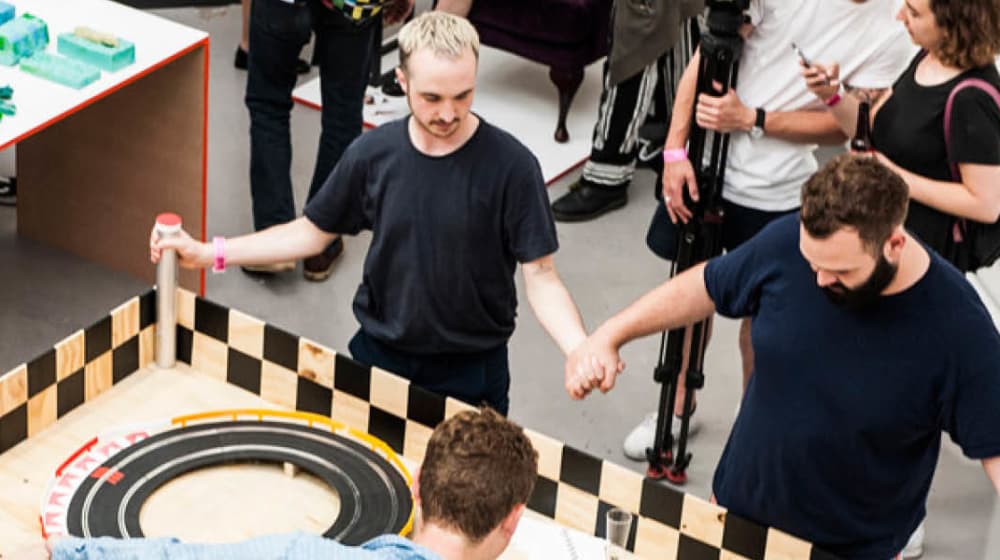 photograph of two men holding hands while powering toy cars by human energy