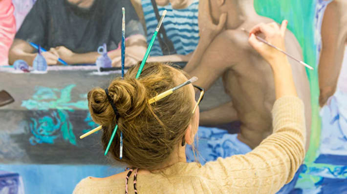 A person with paintbrushes in their hair, painting onto a large canvas