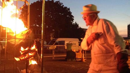 Photograph of man in white clothes and hat standing outside illuminated by a fire at night