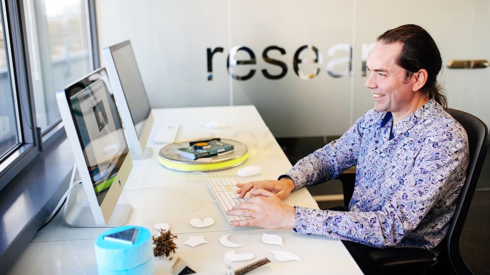 Man in purple shirt smiling whilst sitting at a Mac in a computer lab