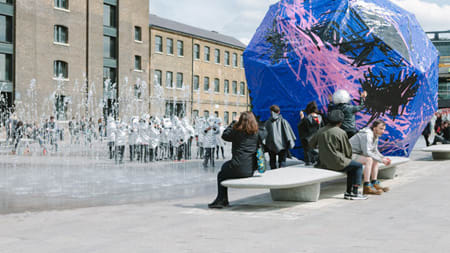 A large purple and blue multi-sided shape is placed in the middle of Granary Square, in front of Central Saint Martins