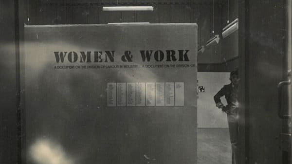 Archive image of a person stood next to an exhibition signs which reads 'Women & Work'