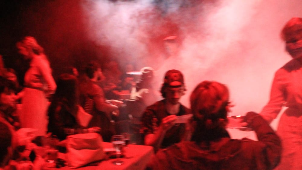 People sittign and standing in a red-lit room full of dry ice