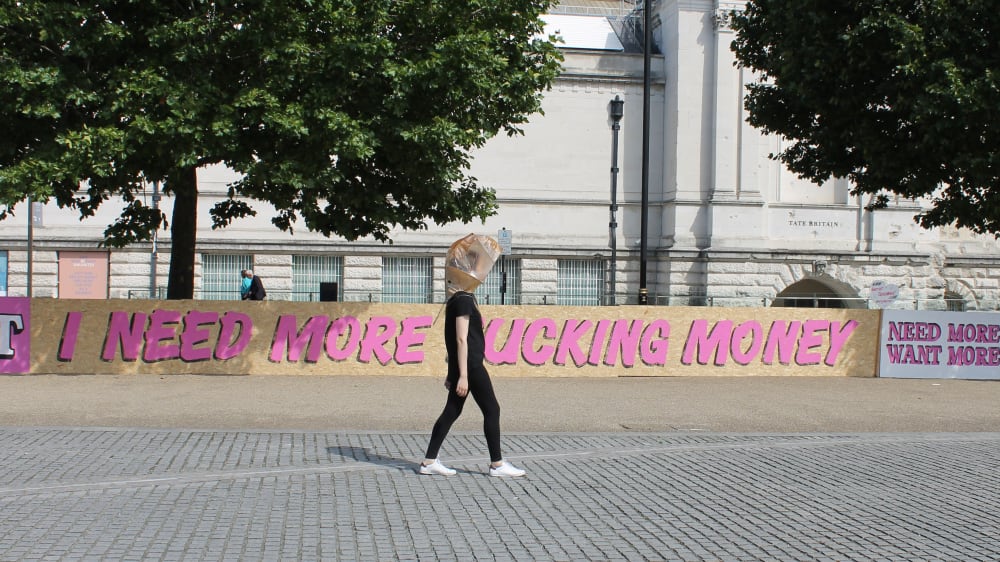 figure wearing a piece of adrienne bennie's design, behind them the fence features hand-written words: 'i need more money'