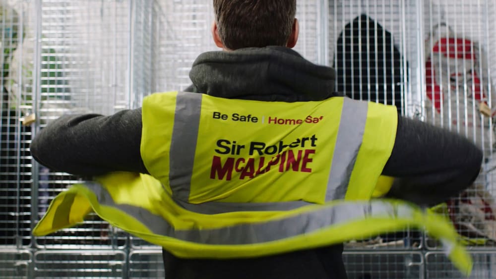Still from a Health and Safety film showing a man facing a white locker, displayer the back of his Sir Robert McAlpine high-vis jacket.
