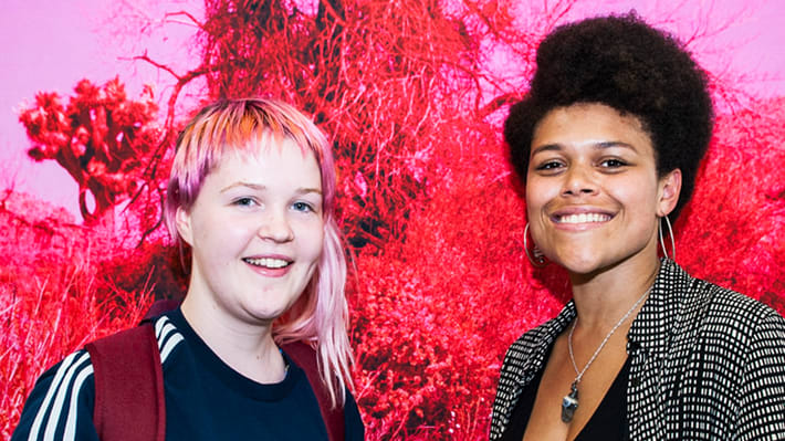 Two women stand in front of a red and pink artwork.