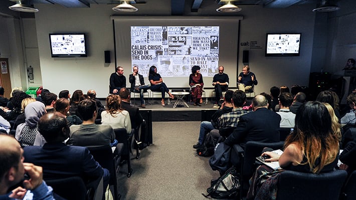A filled conference room featuring a panel of six people on a stage.