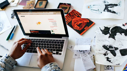view of students hands wprking at a computer with sketches and designs around their work area