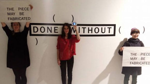 Photo of three woman standing in front of a white wall with 'done without' written on it and holding signs saying