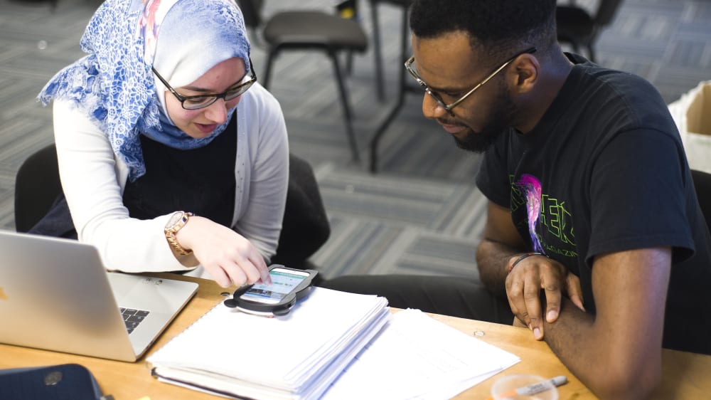 Two students sit and a desk and look at a mobile phone together.