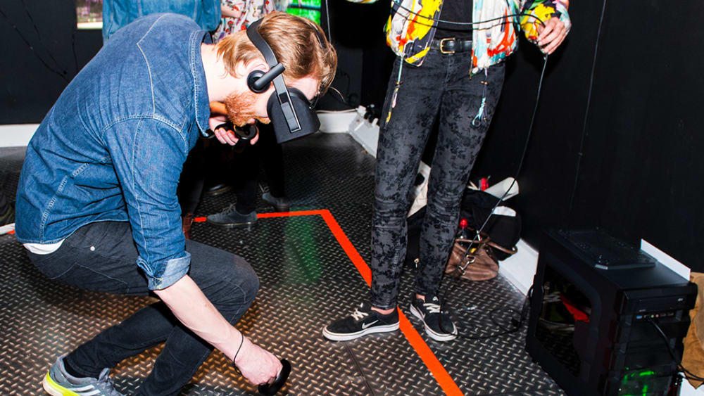 Image of man in denim shirt and jeans wearing a virtual reality headset while looking and bending down within a red rectangle taped to the floor.