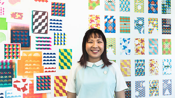 A smiling woman stands in front of a wall display of colourful prints.
