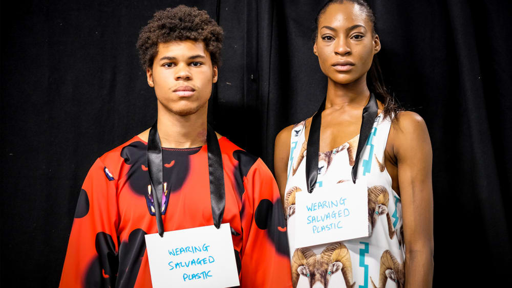 Two models wearing signs saying 'wearing salvaged plastic' from Vin+Omi's LFW presentation