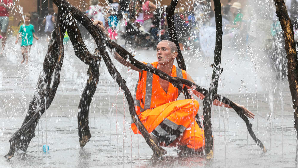A person wearing an orange hi-vis jacket holding a branch in the Granary Square fountains