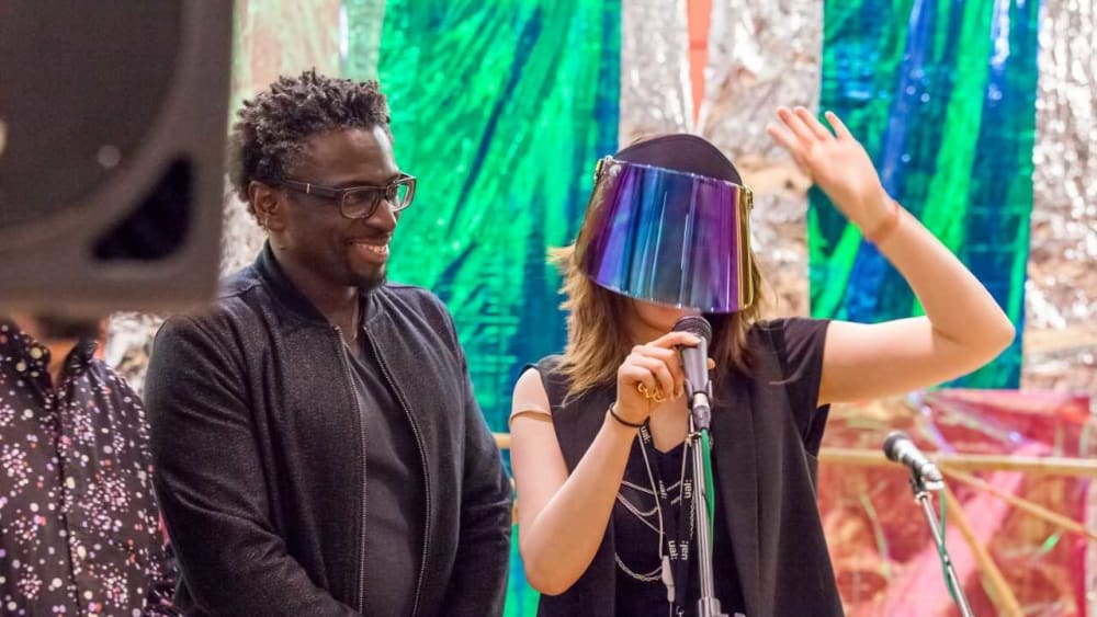 Two people stood in front of a background which is made out of foil. One of the people is wearing a reflective visor over their eyes.