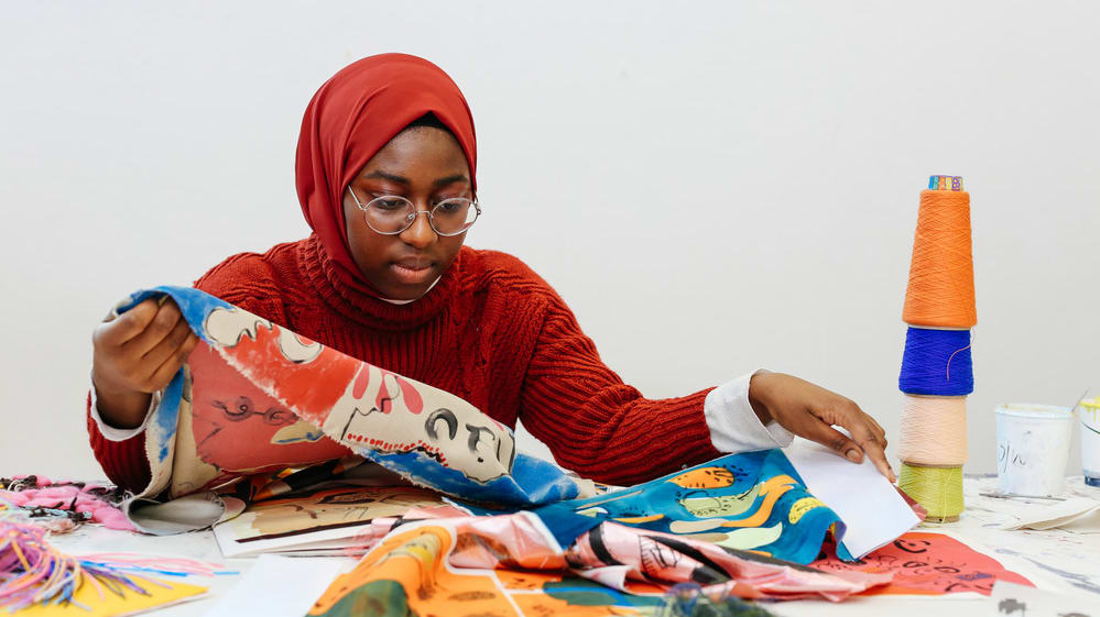 Black female student sitting at workbench looking at various brightly coloured fabrics