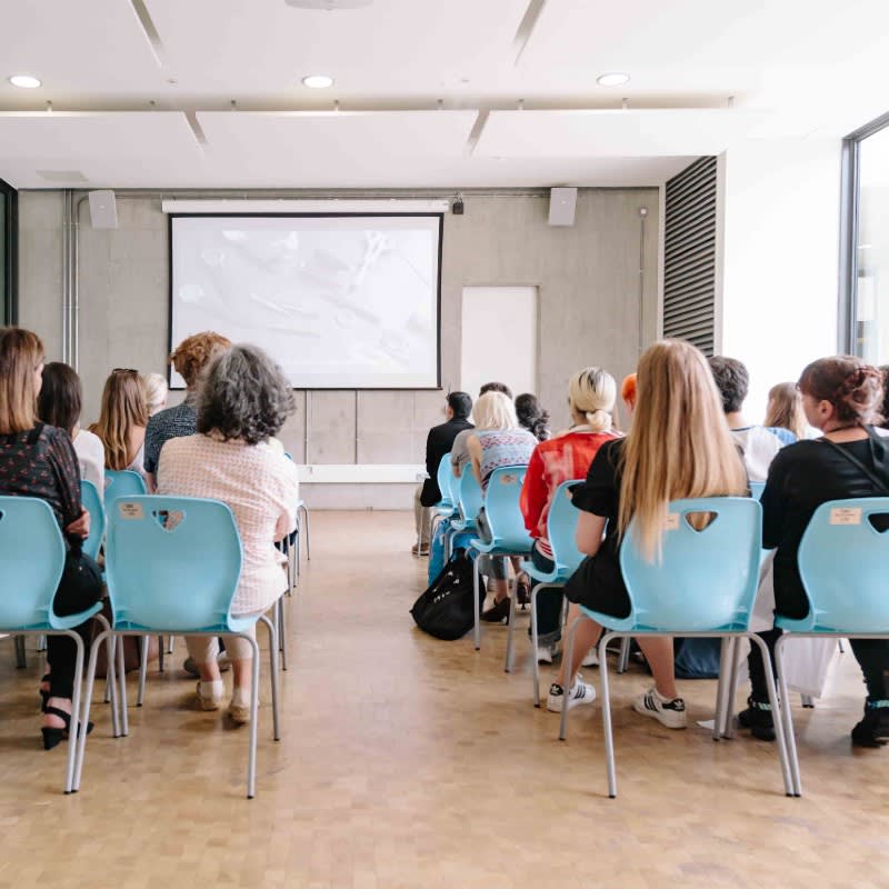 A group of people sit on blue chairs facing a speaker giving a presentation.
