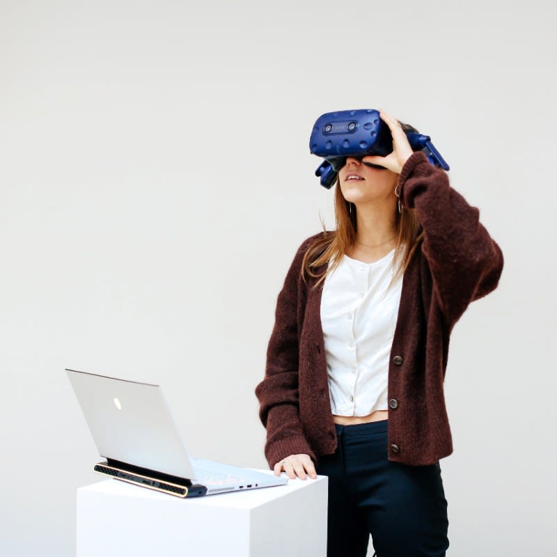 A person wearing virtual reality goggles stands in front of a laptop.