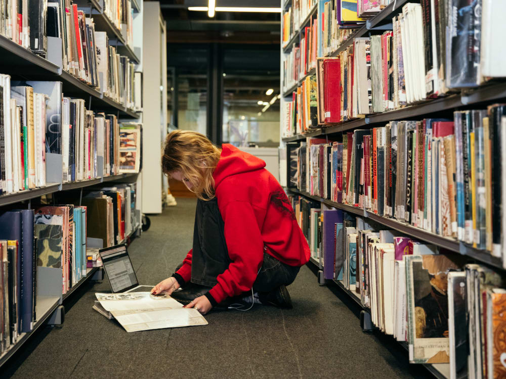 A student in a red hoodie is crouched in the aisle of the library between 2 tall shelf units. She is flicking through an book and has a laptop open beside her.