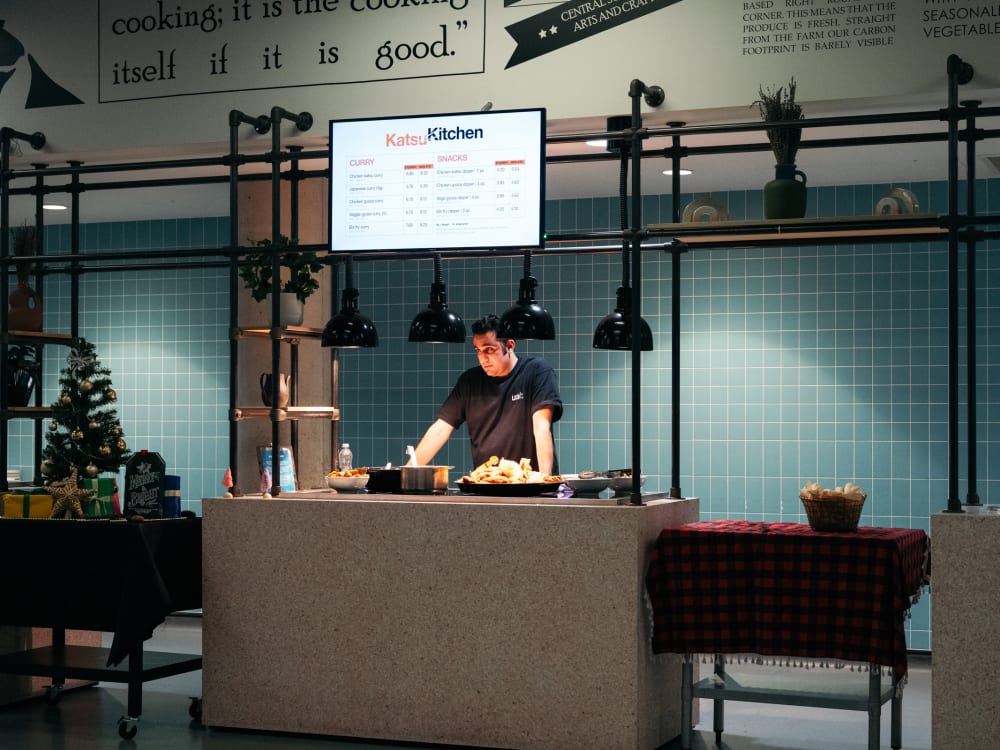 An industrial style canteen counter with a screen above reading 'Katsu Kitchen' and showing a menu. A staff member in a black t-shirt is waiting to serve students and bowls of food are visible on the countertop under heatlamps. The wall behind has square teal tiles and other serving stations are visible to the right and left.
