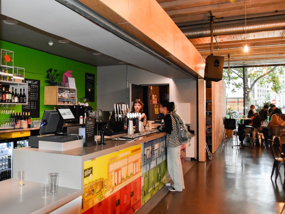 A student is ordering from staff at the bar, which has a brightly coloured branded front. Bar visitors, tables and a window with trees outside are visible. 