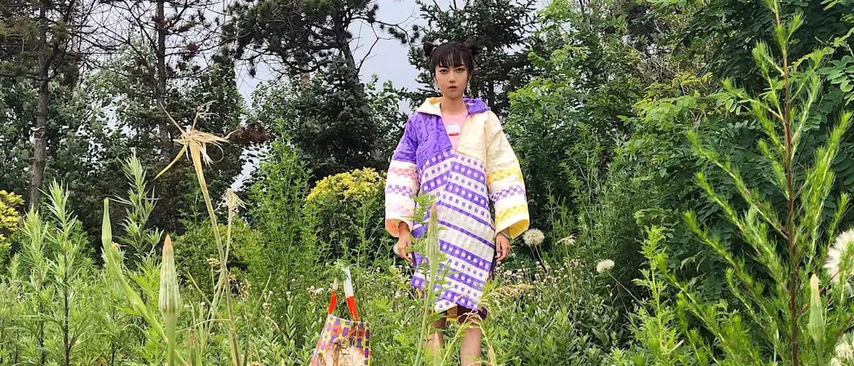 Girl standing amongst grass, tress and bushes wearing a brightly coloured coat.