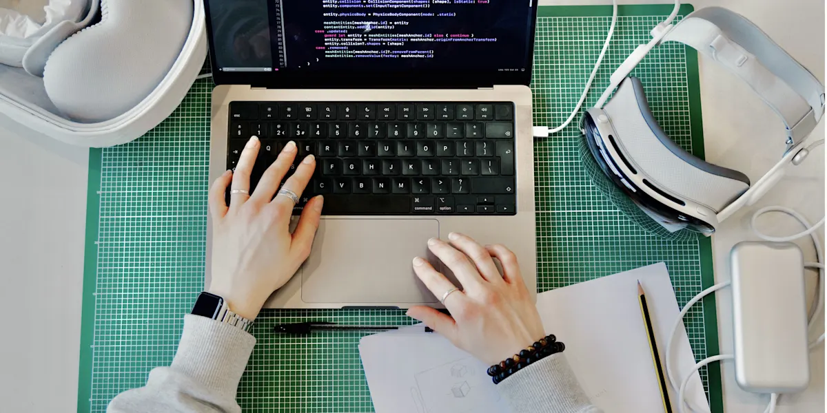 MacBook Pro with colourful Swift code displayed, and an Apple Vision Pro off to the side, on a green cutting mat, with hands on the keyboard.