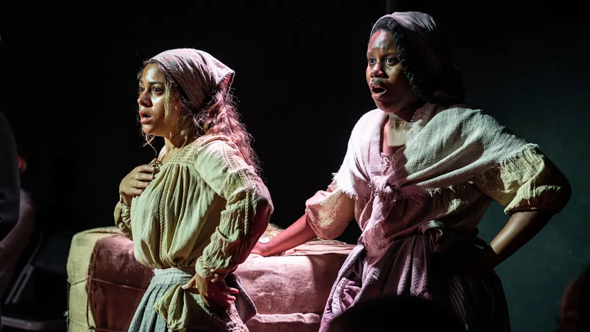 Two performers in period costume stand beside a covered table under dramatic stage lighting.